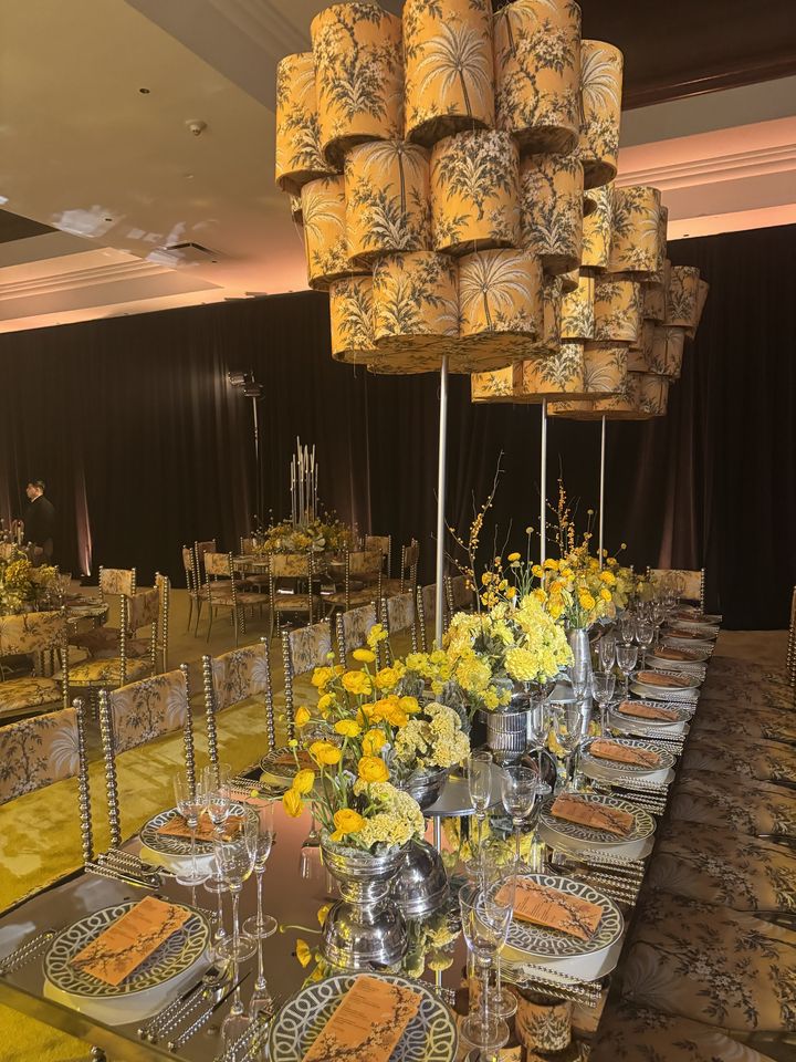 A mirrored long table with yellow ranunculus under palm-print lampshade chandeliers