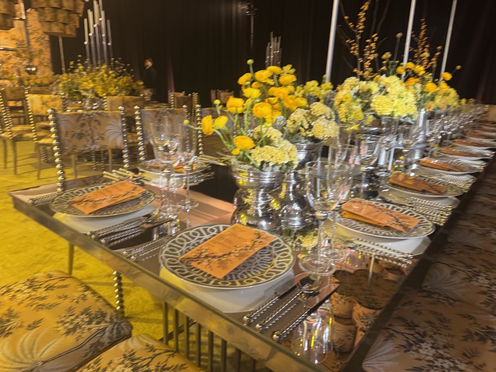 Yellow ranunculus in silver urns on a mirrored table, set with patterned china and gold flatware
